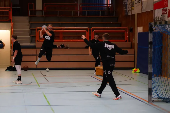 Handballspieler trainieren in einer Sporthalle. Ein Spieler wirft den Ball, während ein anderer sich im Vordergrund zum Wurf vorbereitet.