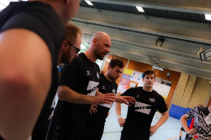 Einige Männer stehen in einem Turnhalle und unterhalten sich. Drei von ihnen tragen schwarze T-Shirts mit dem Aufdruck 