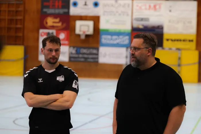 Zwei Männer stehen in einer Sporthalle. Der Mann links trägt ein Handballtrikot mit der Aufschrift „Dragons“, der Mann rechts ein schwarzes T-Shirt. Im Hintergrund sind Werbebanner zu sehen.