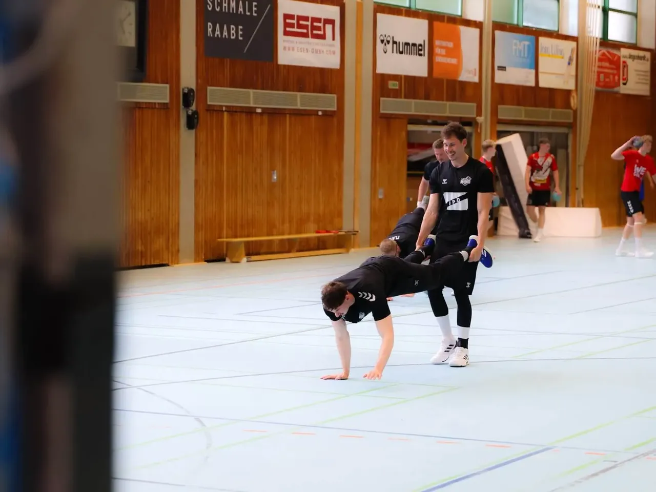 Zwei Männer trainieren eine Kampftechnik in einer Sporthalle. Ein Mann stützt sich auf seine Hände ab, während der andere sein Bein hält. Im Hintergrund sind weitere Sportler zu sehen.