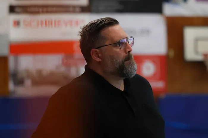 Ein Mann mit Bart und Brille blickt zur Seite, im Hintergrund eine Sporthalle mit Werbebanden.
