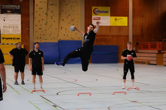 Ein Handballspieler springt in einer Sporthalle in der Luft und wirft den Ball. Im Hintergrund stehen weitere Spieler und Trainingsgeräte.