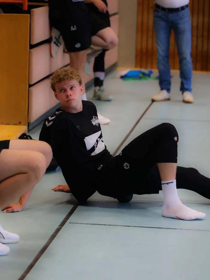 Ein junger Handballspieler mit lockigem blonden Haar sitzt auf der Turnhallenmatte und blickt nachdenklich in die Ferne. Im Hintergrund sind weitere Sportler zu sehen.