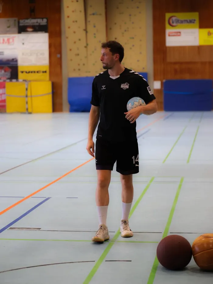 Ein Handballspieler in schwarzer Sportkleidung steht in einer Sporthalle und hält einen Handball. Im Hintergrund sind weitere Sportgeräte und Werbebanner zu sehen.