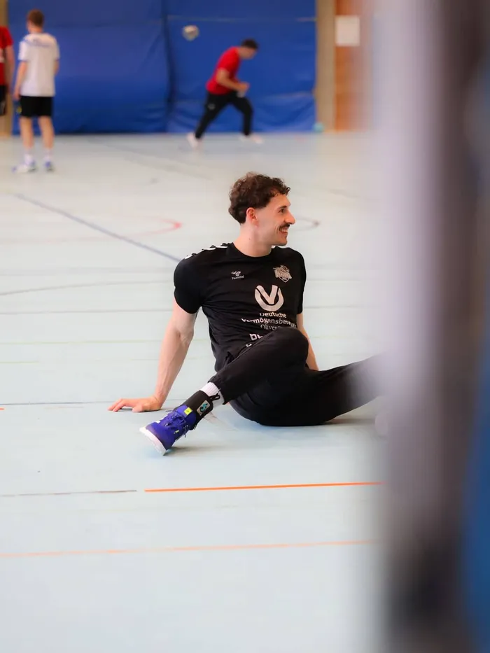 Ein Handballspieler sitzt auf dem Hallenboden und dehnt seine Beine. Im Hintergrund sind weitere Spieler beim Training zu sehen.