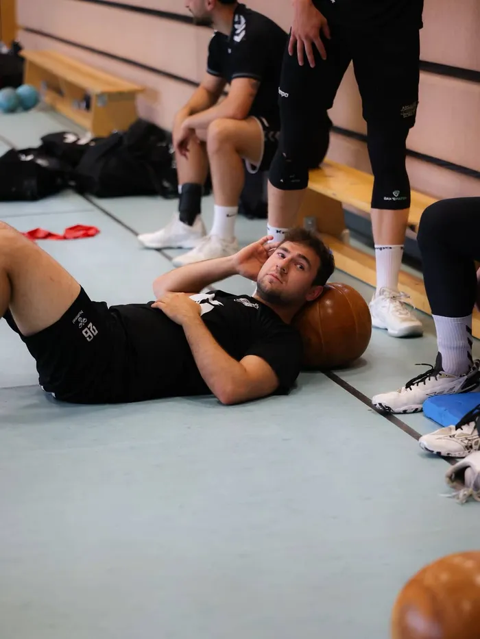 Ein Handballspieler macht Bauchmuskelübungen auf einer Matte in einer Sporthalle, während Teamkollegen im Hintergrund sitzen.