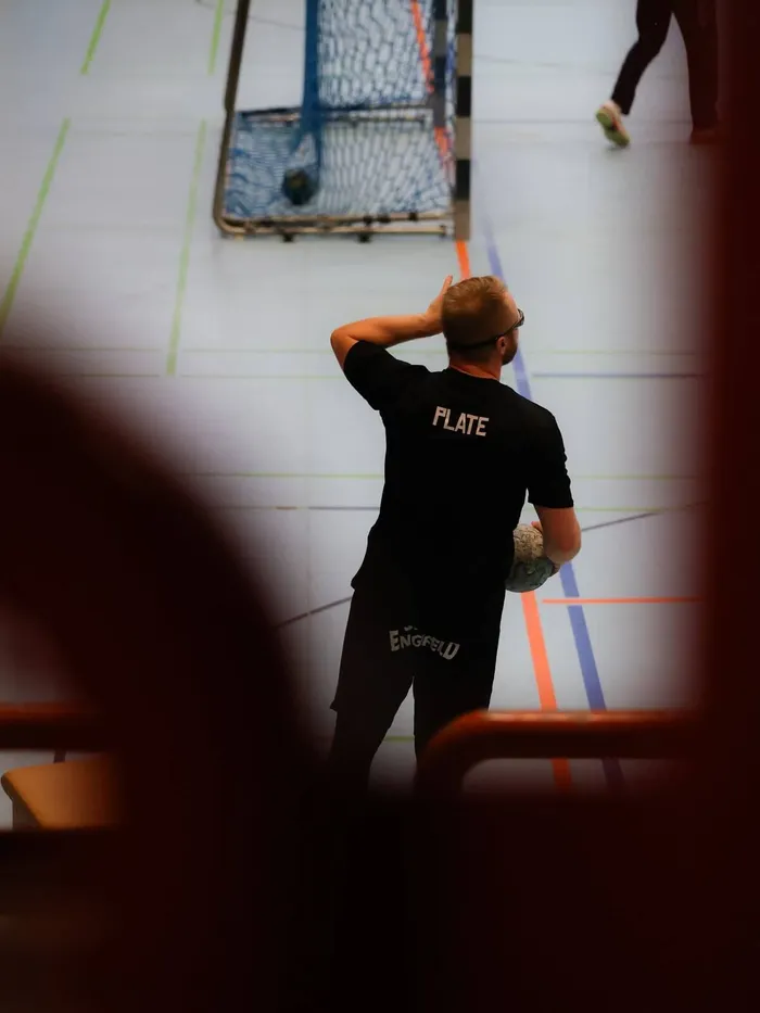 Ein Handballtrainer steht mit dem Rücken zum Betrachter auf der Halle. Im Hintergrund ein Handballtor.