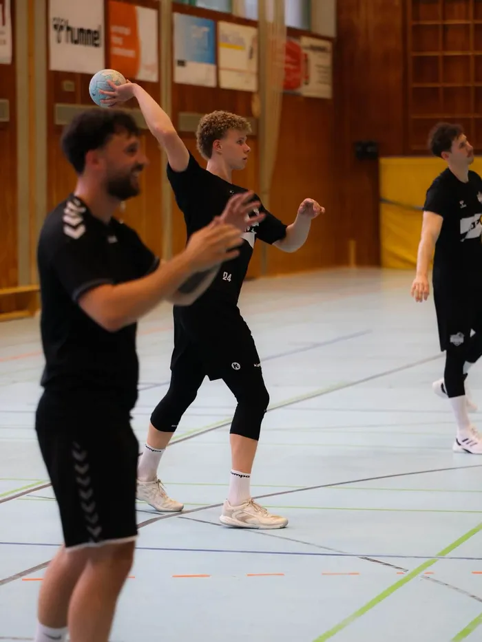 Ein Handballspieler wirft den Ball in einer Sporthalle. Im Hintergrund sind weitere Spieler zu sehen.