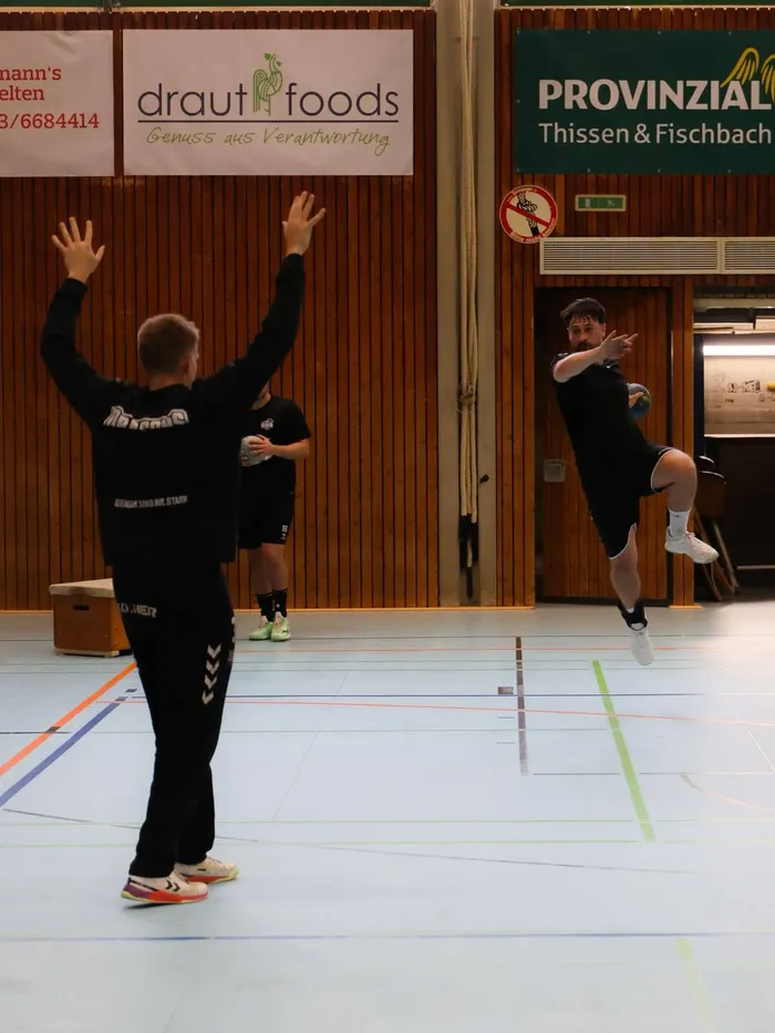 Ein Handballspieler springt in der Luft, während ein zweiter Spieler mit erhobenen Armen darunter steht. Im Hintergrund sind Werbebanner und eine Holzwand zu sehen.
