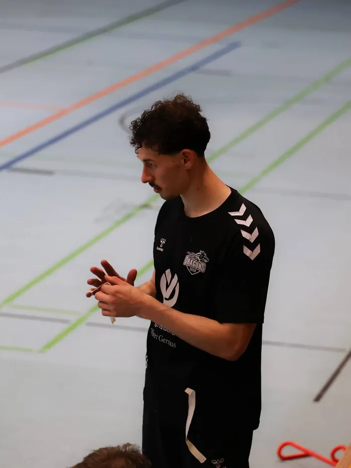 Ein Mann mit lockigem Haar gestikuliert mit den Händen, während er ein schwarzes Trikot mit dem Aufdruck 