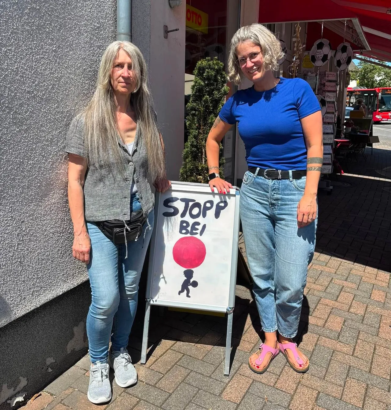 Zwei Frauen stehen neben einem weißen Aufsteller mit der Aufschrift „STOPP BEI“ und einem roten Kreis im Rahmen einer Plakataktion.