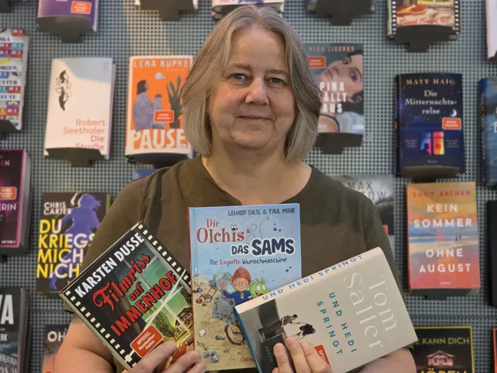 Eine Frau hält die Bücher „Filmriss und Immenhof“, „Die Olchis & Das Sams“ und „Und Hedi springt“ vor einer Wand voller Bücher.