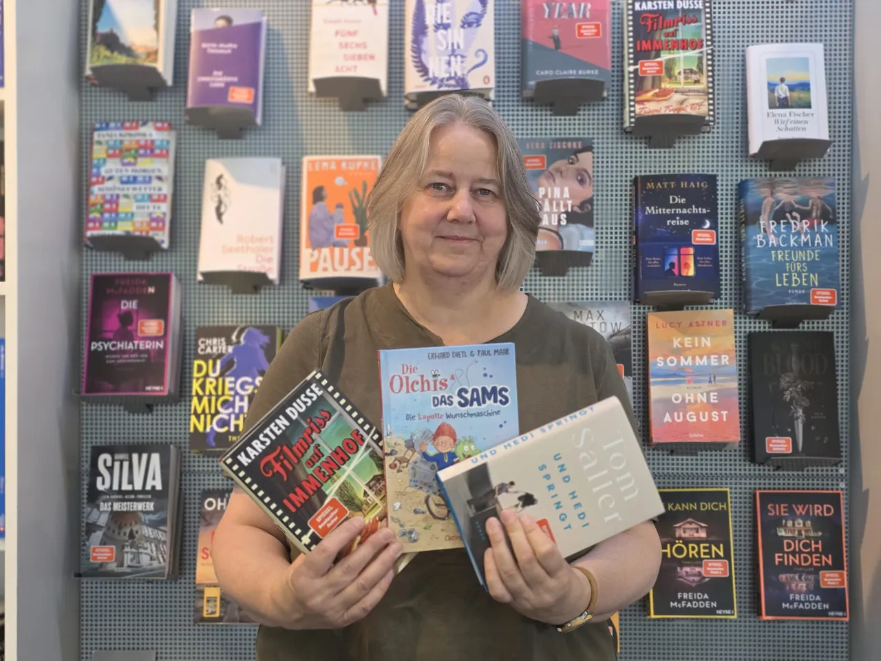 Eine Frau hält drei Bücher vor einer Wand mit zahlreichen Büchern in einem Display.