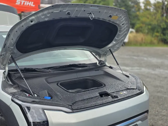 Geöffnete Motorhaube eines silbernen Autos mit einer glatten, schwarzen Abdeckung im Motorraum.
