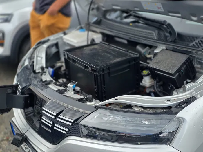 Blick in den geöffneten Motorraum eines silbernen Autos mit technischen Komponenten und einer großen schwarzen Box.
