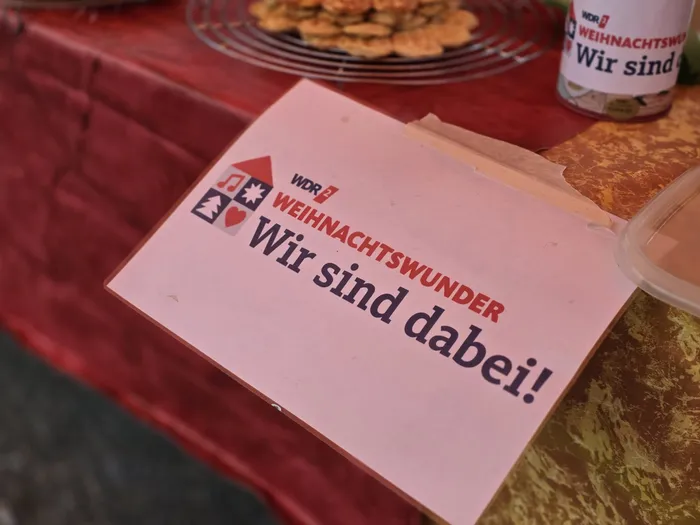 Ein Schild mit der Aufschrift „WDR 2 WEIHNACHTSWUNDER Wir sind dabei!“ liegt auf einem Tisch neben Gebäck.