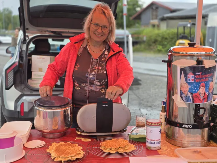 Eine lächelnde Frau in einer roten Jacke steht an einem Tisch mit Waffeln und einem Getränkespender vor einem weißen Auto.