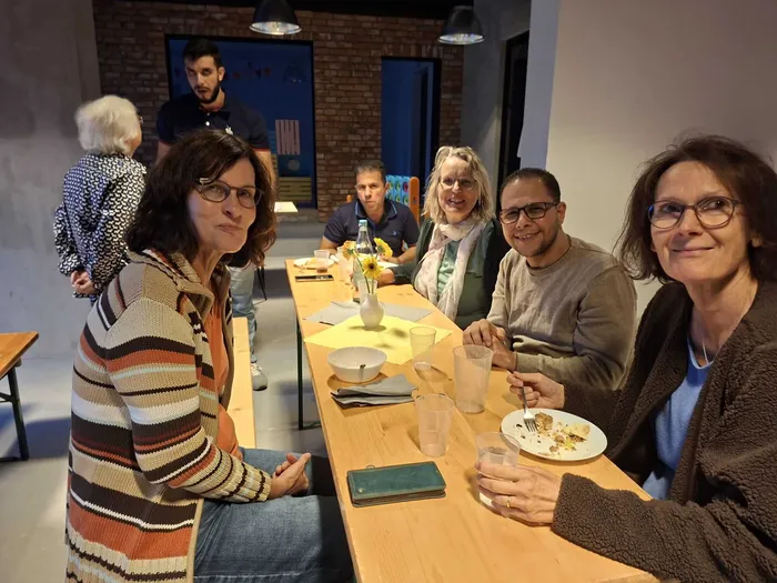 Eine Gruppe von Menschen sitzt lächelnd an einem langen Holztisch mit Speisen und Getränken bei einem Kultur-Häppchen-Fest.