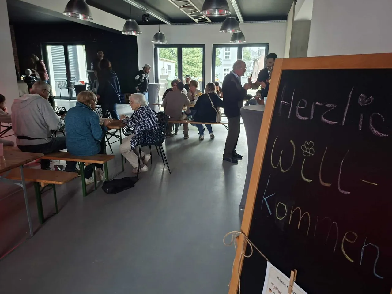 Eine Tafel mit der Aufschrift „Herzlich Willkommen“ steht im Vordergrund eines Raumes, in dem Menschen an Tischen zusammensitzen.