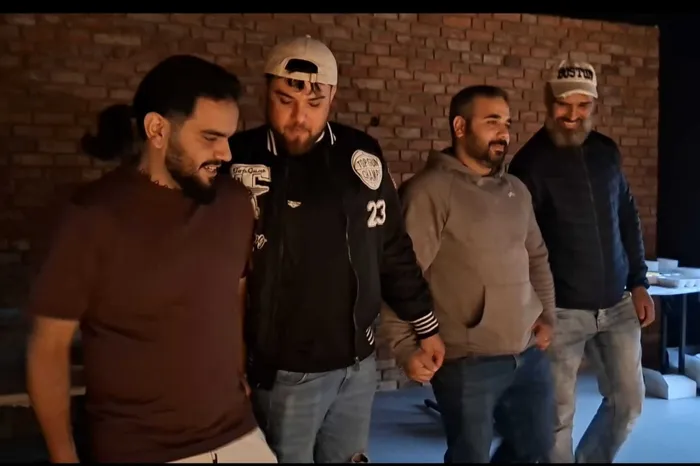 Vier Männer stehen eng beieinander vor einer Ziegelwand.