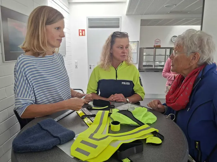 Eine Polizistin und zwei Frauen sitzen an einem Tisch mit gelben Sicherheitswesten.