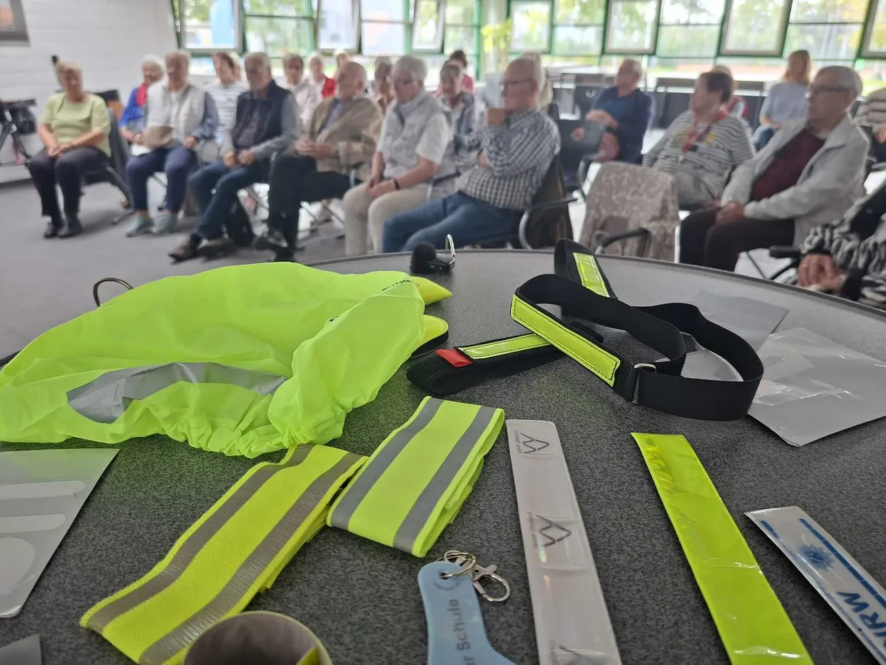 Neonfarbene Warnwesten und Reflektoren liegen auf einem Tisch vor einem Publikum bei einem Vortrag zur Europäischen Mobilitätswoche.