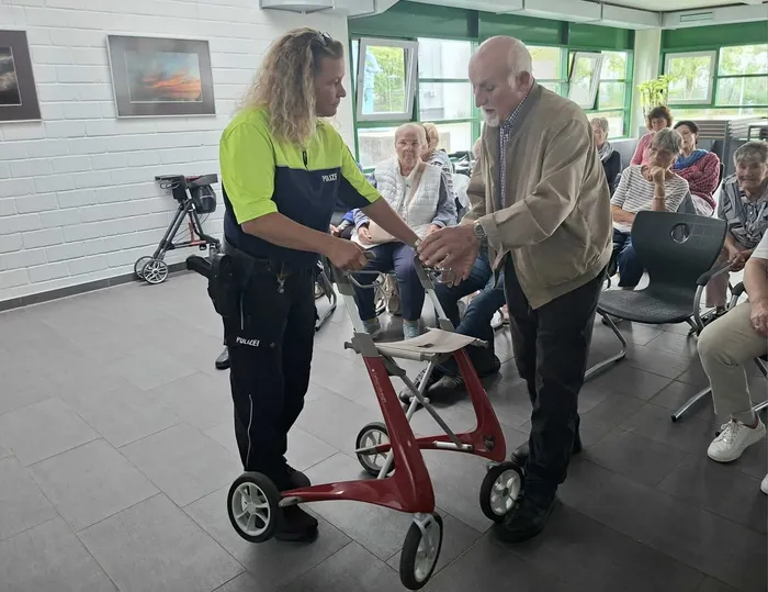 Eine Polizistin und ein älterer Mann halten gemeinsam einen roten Rollator vor einem Publikum in einem Innenraum.