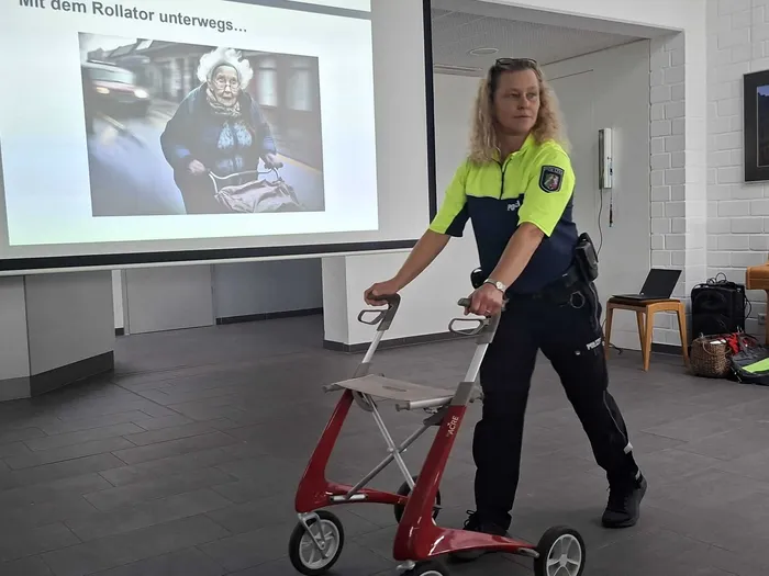 Eine Polizistin schiebt einen roten Rollator vor einer Projektionsfläche, die eine ältere Person mit Rollator zeigt.