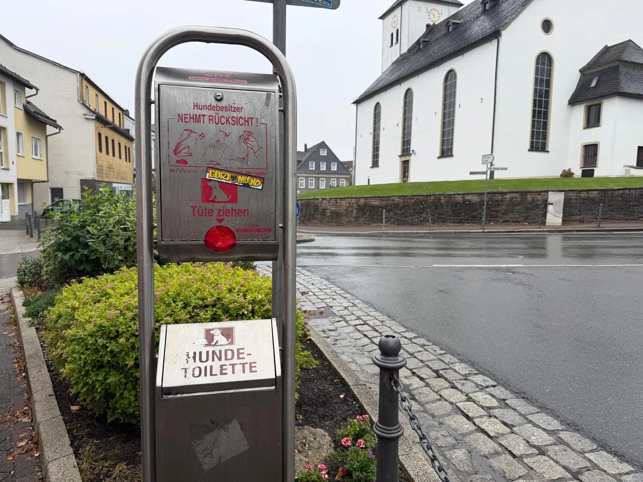 Metallener Spender für Hundekotbeutel mit Aufklebern und der Aufschrift „HUNDE-TOILETTE“ an einem Gehweg.