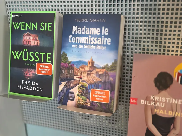 Drei Bücher in einem Metallregal: „Wenn sie wüsste“ von Freida McFadden, „Madame le Commissaire und die tödliche Rallye“ von Pierre Martin und ein Buch von Kristine Bilkau.