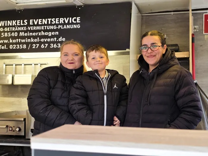 Drei Personen stehen vor einem Imbisswagen mit der Aufschrift „Kattwinkel Eventservice“. Links eine Frau, in der Mitte ein Junge und rechts eine weitere Frau, alle mit Winterjacken bekleidet.