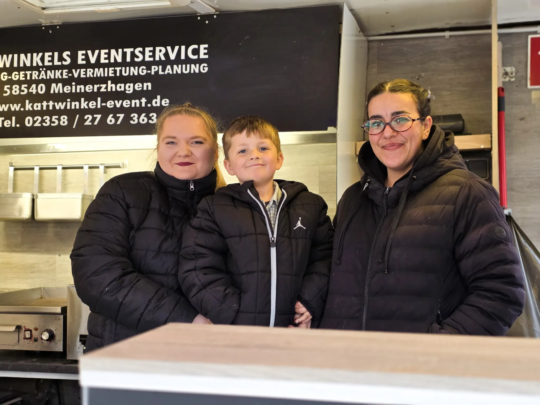 Drei Personen stehen vor einem Imbisswagen mit der Aufschrift „Kattwinkel Eventservice“. Links eine Frau, in der Mitte ein Junge und rechts eine weitere Frau, alle mit Winterjacken bekleidet.