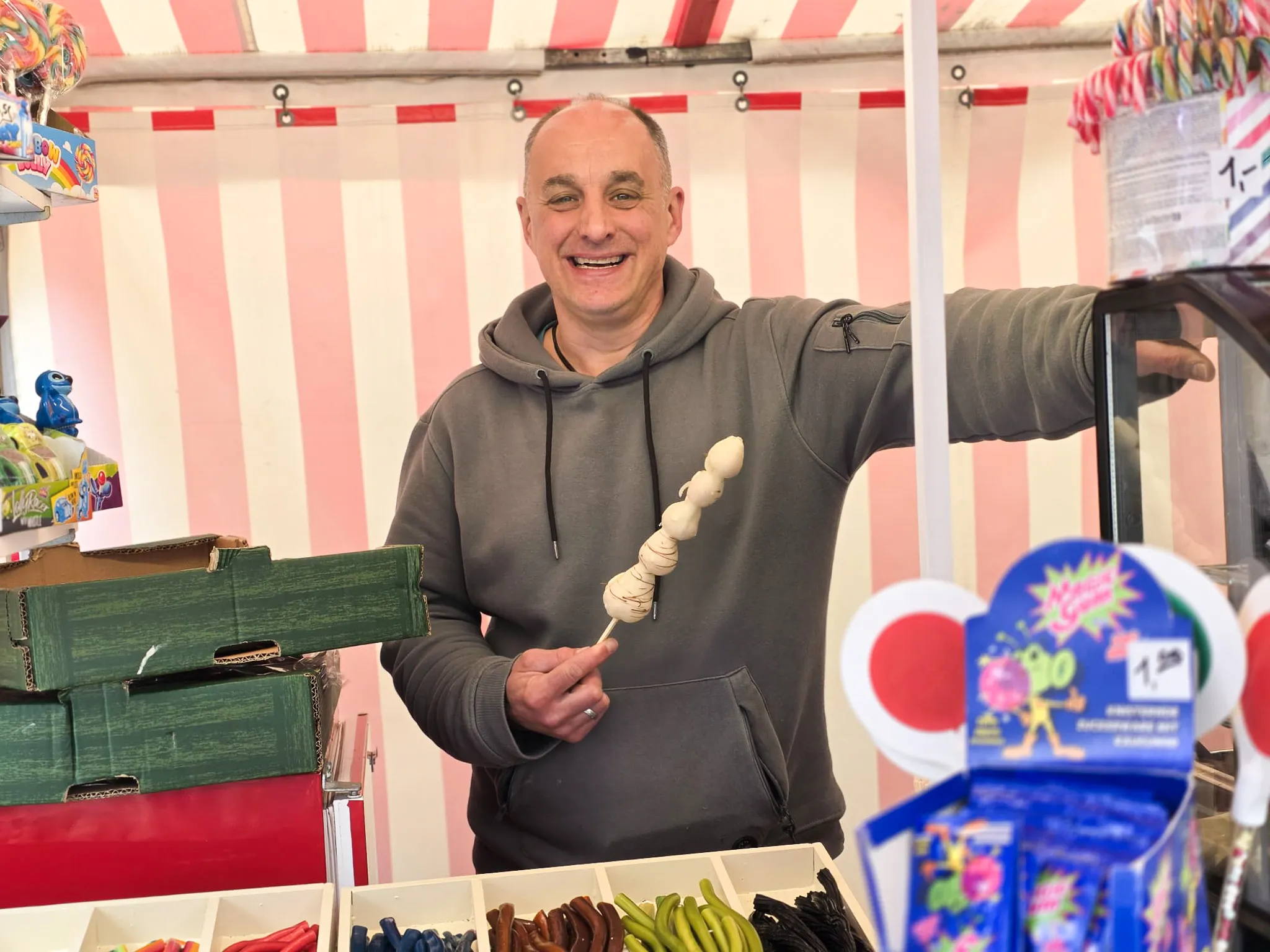 Ein Mann mittleren Alters lächelt in einem Süßigkeitenstand und hält eine Spieß mit weißen Süßigkeiten hoch.