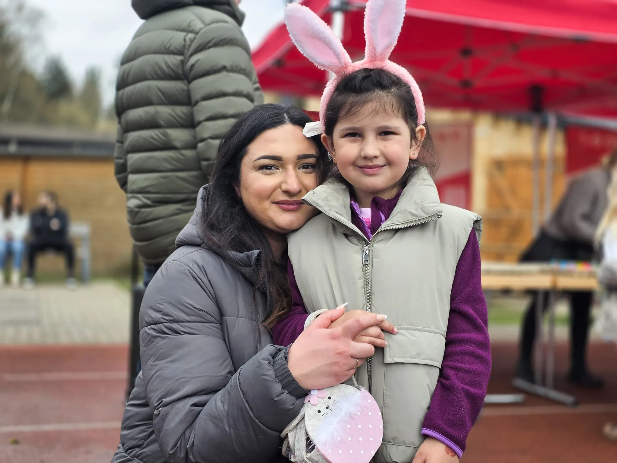 Eine Frau umarmt ein Mädchen mit Hasenohren auf dem Kopf. Im Hintergrund sind weitere Menschen und ein rotes Zelt zu sehen.
