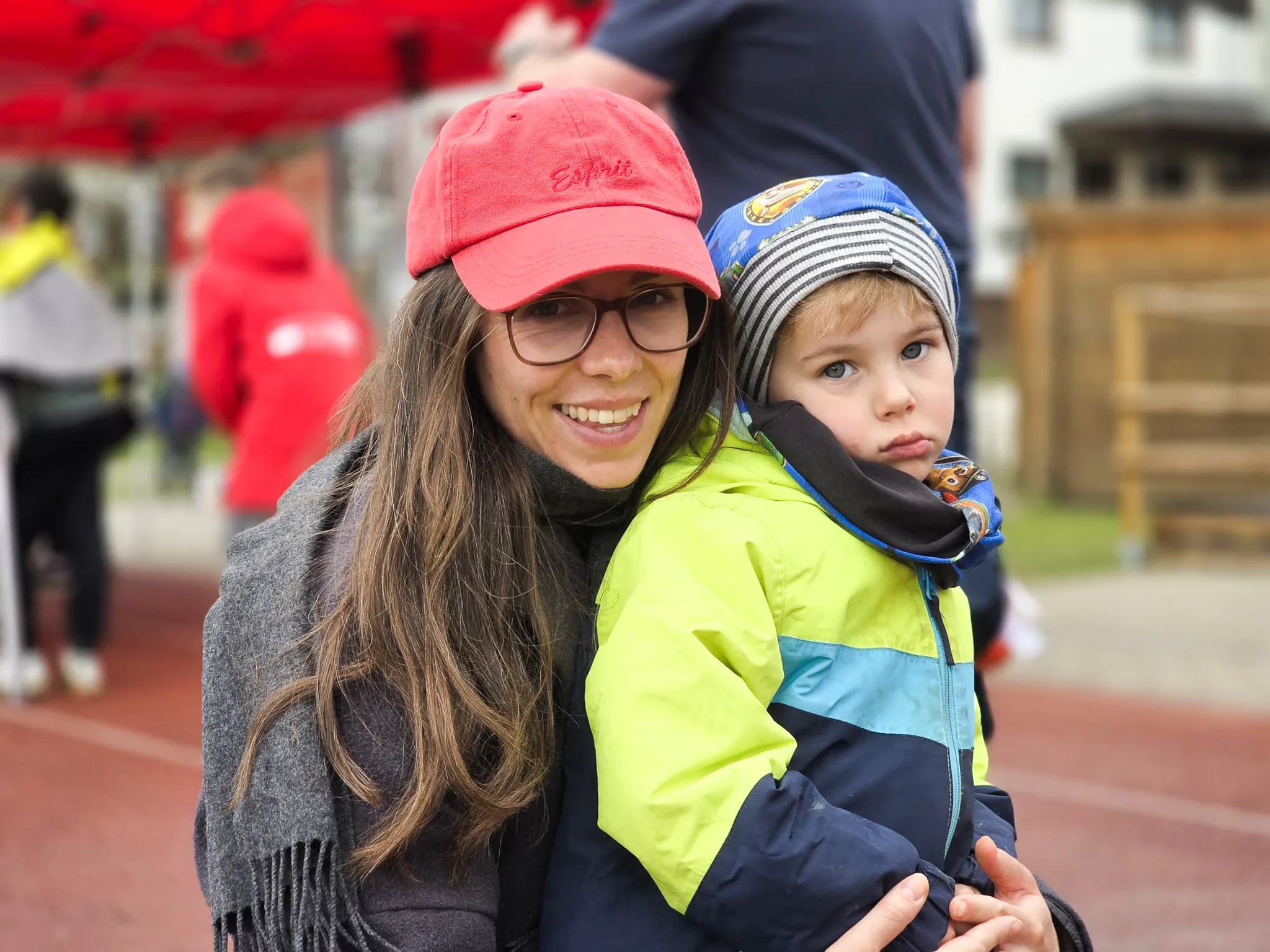 Eine junge Frau mit roter Baseballkappe hält ein Kleinkind im Arm, beide schauen in die Kamera. Im Hintergrund sind weitere Menschen und ein roter Pavillon zu sehen.
