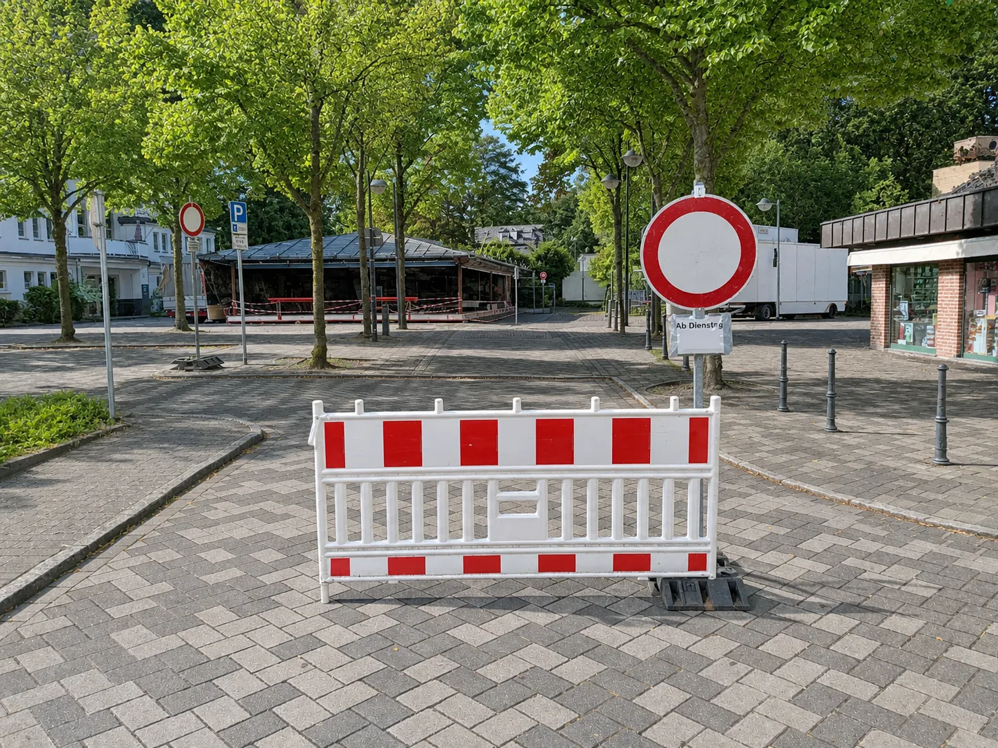Rote und weiße Absperrgitter versperren den Zugang zu einem Platz, im Hintergrund ein Verbotsschild für Fahrzeuge und ein überdachter Bereich.