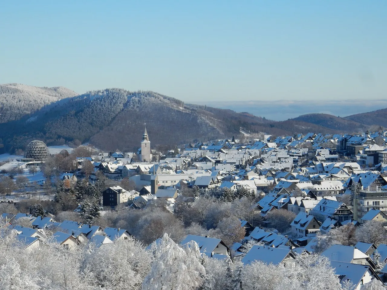 Schneebedeckte Dächer von Häusern und ein Kirchturm prägen das Stadtbild von Winterberg, im Hintergrund bewaldete Hügel.