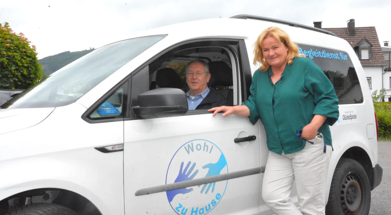 Ein Mann sitzt lächelnd am Steuer eines weißen VW Caddy, während eine Frau neben dem Fahrzeug steht und ihre Hand an die offene Tür legt. Auf der Tür ist ein blauer Kreis mit einer weißen Hand und dem Text „Wohl zu Hause“ abgebildet.