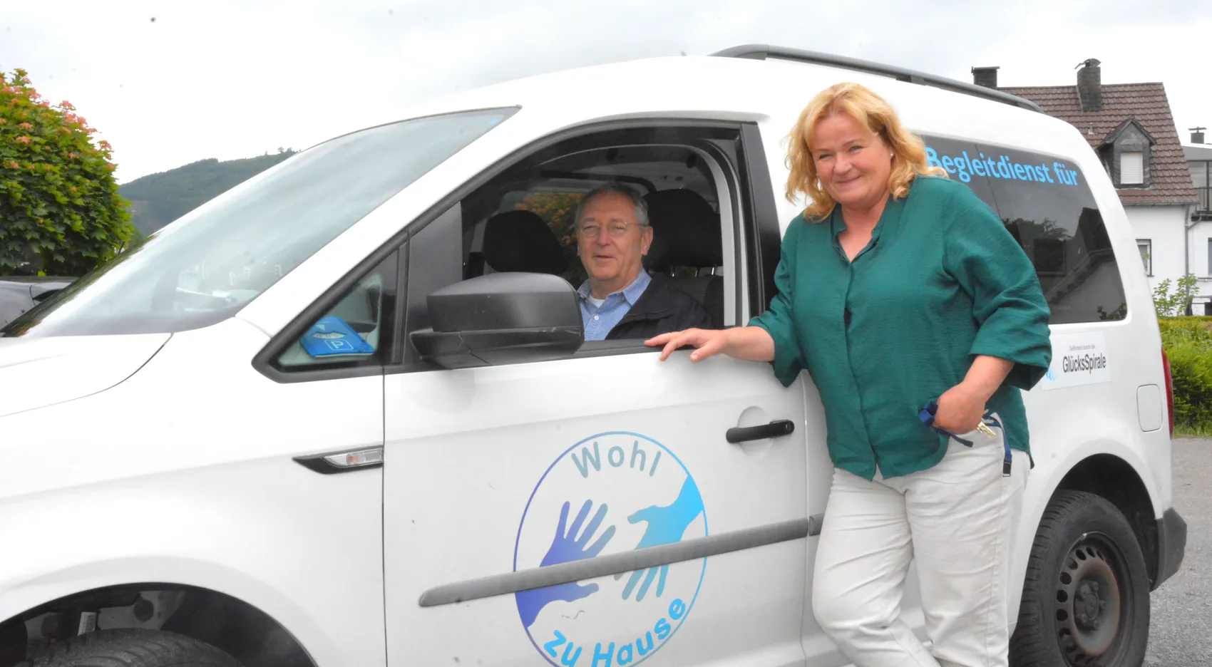Ein Mann sitzt lächelnd am Steuer eines weißen VW Caddy, während eine Frau neben dem Fahrzeug steht und ihre Hand an die offene Tür legt. Auf der Tür ist ein blauer Kreis mit einer weißen Hand und dem Text „Wohl zu Hause“ abgebildet.