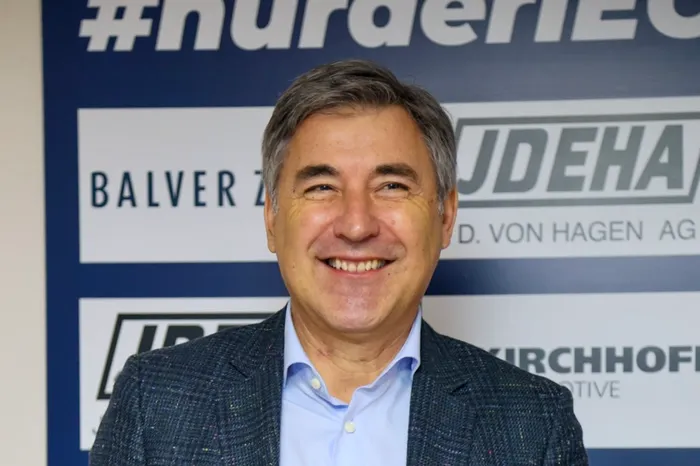 Wolfgang Brück, lächelnd in Anzug vor blauem Hintergrund mit Werbeaufdruck, tritt als Geschäftsführer der Iserlohn Roosters zurück.