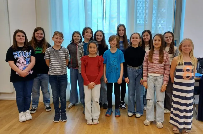 Eine Gruppe von etwa zwölf Kindern und Jugendlichen steht lächelnd in einer Reihe vor einem Fenster. Sie tragen Alltagskleidung, darunter Jeans, T-Shirts und Kleider.