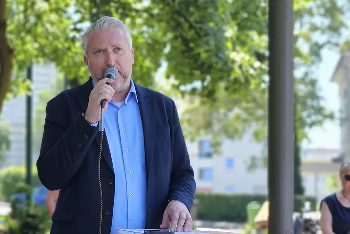 Ein Mann mit blonden Haaren und Bart hält ein Mikrofon in der Hand und spricht. Er trägt einen dunklen Anzug und ein hellblaues Hemd. Im Hintergrund sind Bäume und Gebäude zu sehen.