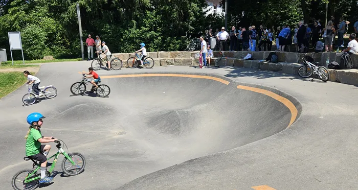 Kinder fahren mit Fahrrädern in einem Skatepark, während im Hintergrund Zuschauer stehen.