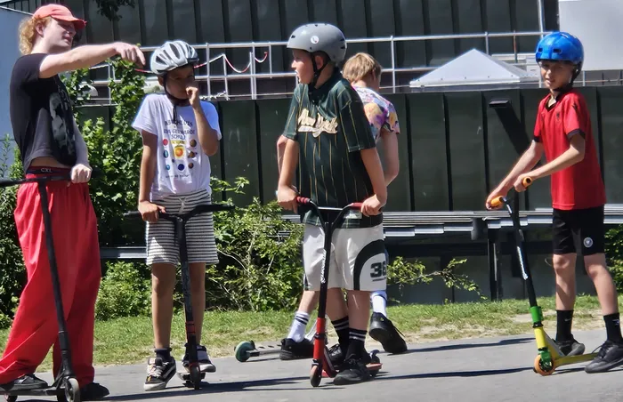 Fünf Kinder und Jugendliche fahren auf Rollern durch eine Fußgängerzone. Sie tragen Helme und sommerliche Kleidung. Im Hintergrund sind Gebäude und Bäume zu sehen.