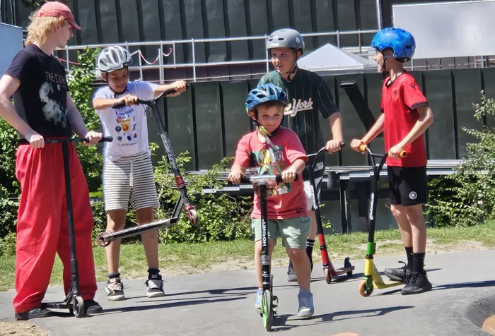 Fünf Kinder und Jugendliche fahren auf Rollern und einem Tretroller auf einem Platz vor einem modernen Gebäude. Alle tragen Helme und sind in Bewegung.