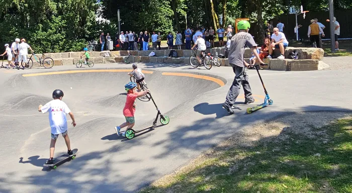 Kinder und Jugendliche fahren auf Skateboards und Scootern in einem Skatepark, während im Hintergrund Zuschauer stehen.