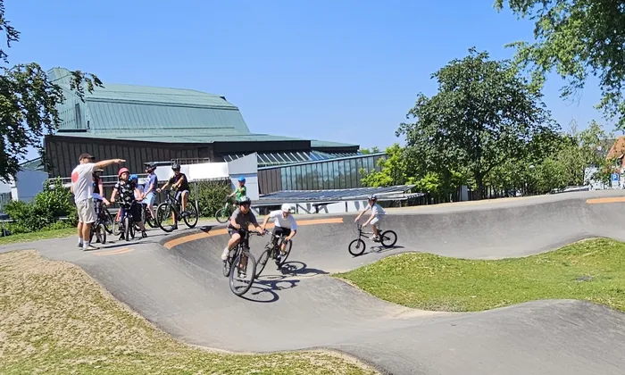 Mehrere Kinder und Jugendliche fahren mit ihren Fahrrädern in einem Skatepark. Im Hintergrund steht ein modernes Gebäude mit einem grünen Dach. Ein Mann zeigt mit dem Finger in eine Richtung.