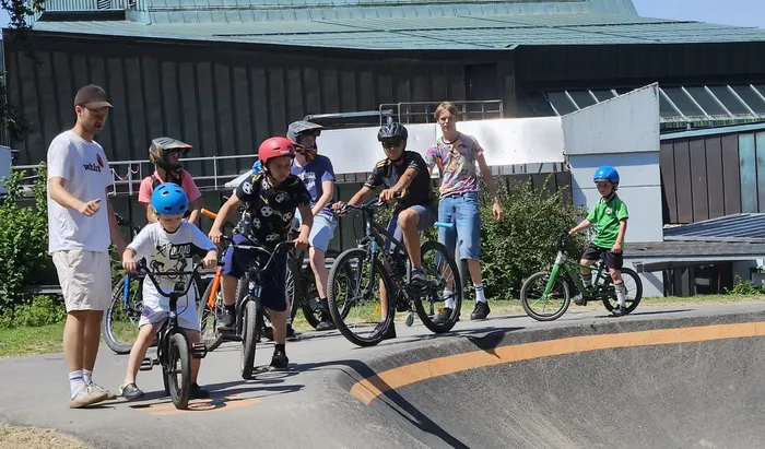 Eine Gruppe Kinder und Jugendliche übt mit Fahrrädern auf einer kleinen Rampe vor einem modernen Gebäude. Einige tragen Helme und sportliche Kleidung.