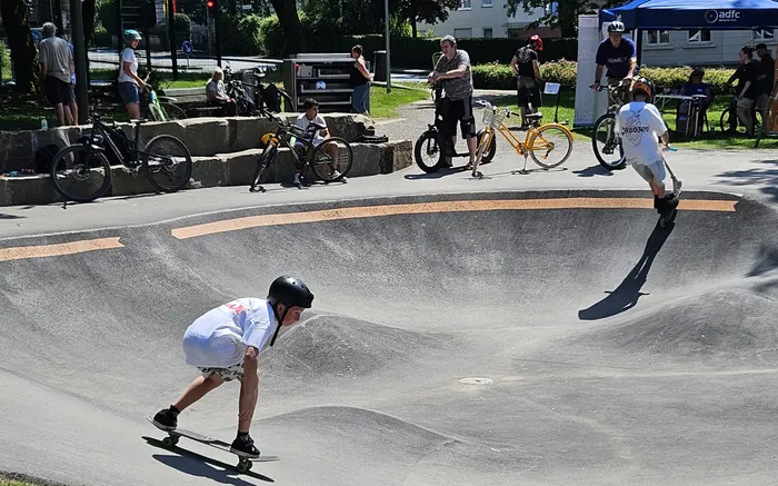 Zwei Jugendliche fahren auf Skateboards in einer Beton-Skateanlage, während im Hintergrund Zuschauer stehen und Fahrräder geparkt sind.