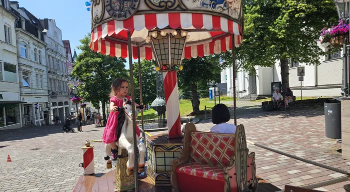 Ein kleines Mädchen sitzt auf einem bunten, beweglichen Pferd, während eine andere Person auf einem Sitz gegenüber sitzt. Im Hintergrund eine belebte Stadtansicht mit Geschäften und Passanten.
